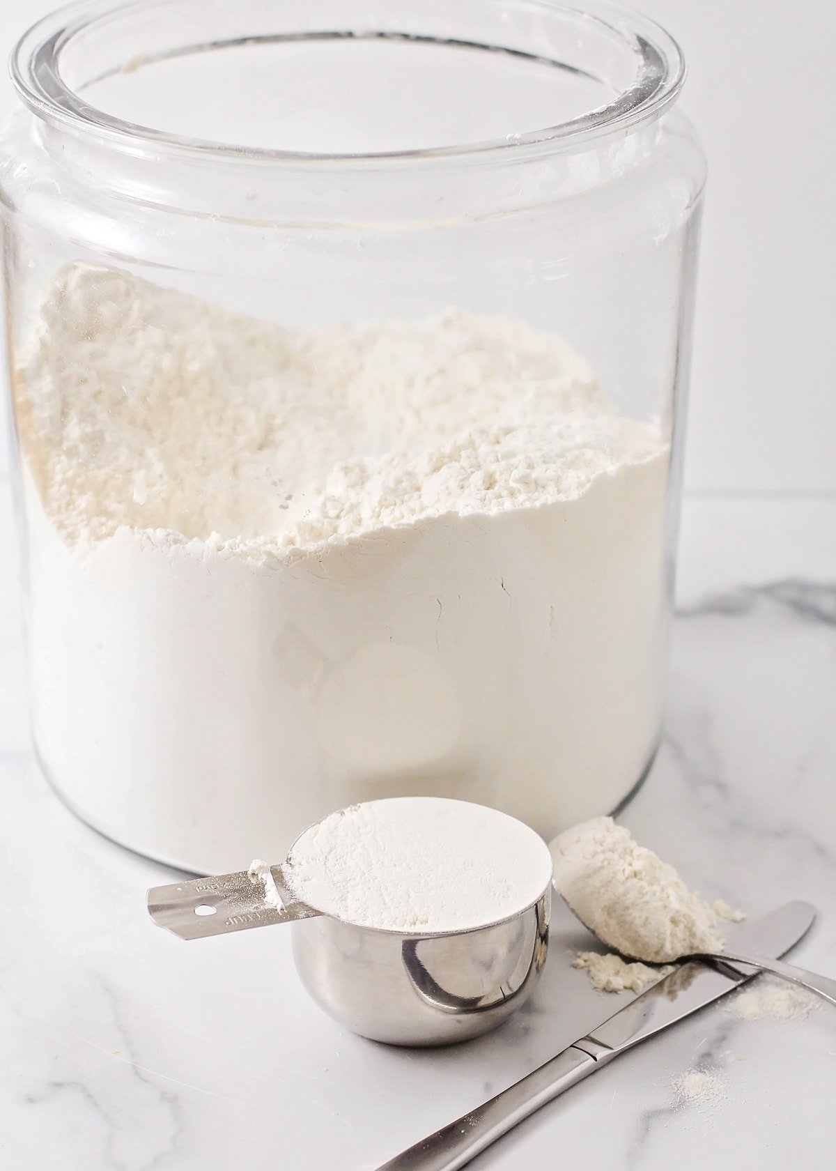 A measuring cup filled with flour in front of a glass jar.